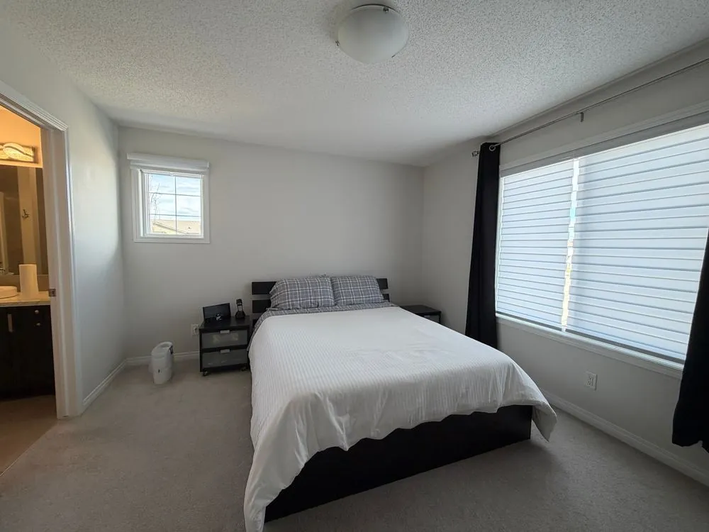 Bedroom before staging: plain bedding, bare walls, cluttered nightstand and window blinds half closed